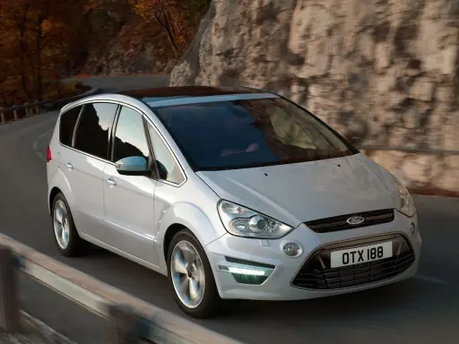 Biały Ford S-Max 2.0 TDCI pokonuje zakręt na malowniczej drodze wśród jesiennych drzew i skał.