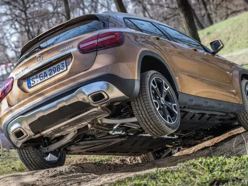 Złoty Mercedes GLA 220 4MATIC pokonuje nierówny teren, unosząc jedno koło.