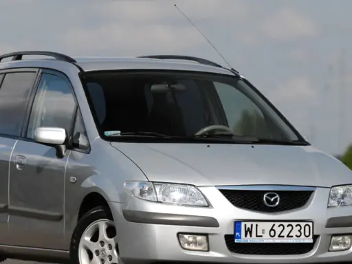 Srebrna Mazda Premacy z relingami dachowymi, zaparkowana na tle nieba i pól.