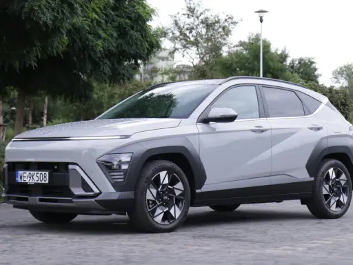 Nowy Hyundai Kona Hybrid w kolorze jasnoszarym, zaparkowany na brukowanej nawierzchni, otoczony zielenią.