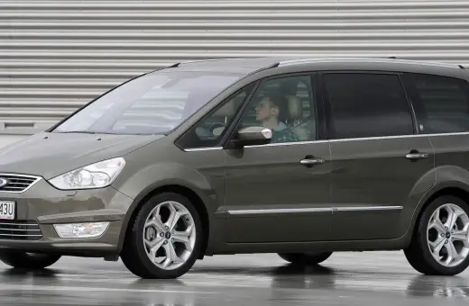 Szary Ford Galaxy MK2 z kierowcą za kierownicą, zaparkowany na mokrej nawierzchni przed budynkiem z poziomymi żaluzjami.