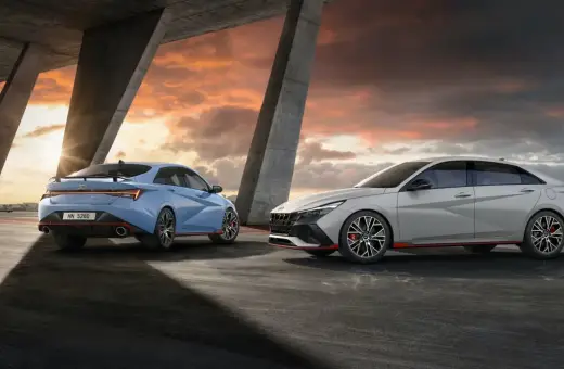 Dwa sportowe Hyundai Elantra N, jeden niebieski, drugi szary, stoją pod betonowym zadaszeniem o zachodzie słońca.