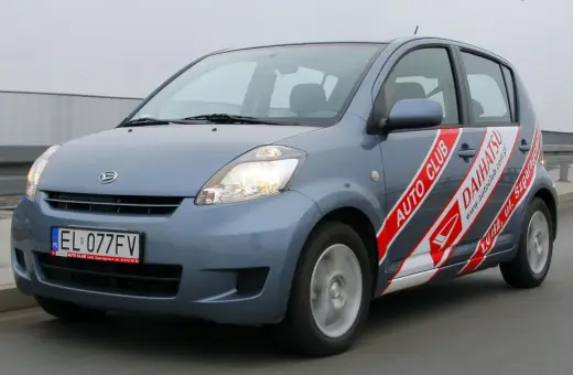 Niebieski Daihatsu Sirion z naklejkami "Auto Club Daihatsu" na drzwiach, poruszający się po drodze.