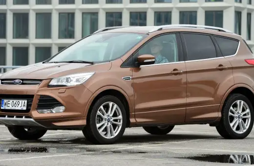 Brązowy Ford Kuga 2017 z kierowcą w okularach, zaparkowany na mokrym parkingu przed nowoczesnym budynkiem.
