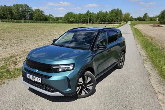 Nowy Opel Frontera 2025 w turkusowym kolorze na wiejskiej drodze, otoczony polami i drzewami.