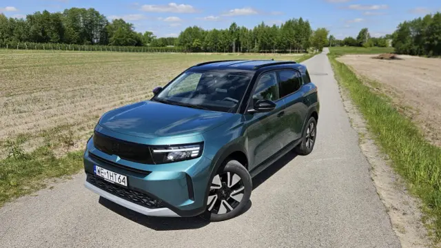 Nowy Opel Frontera 2025 w turkusowym kolorze na wiejskiej drodze, otoczony polami i drzewami.