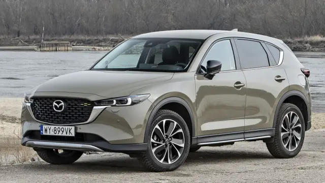Nowa Mazda CX-5 w kolorze piaskowym, zaparkowana nad brzegiem rzeki, gotowa na przygodę.