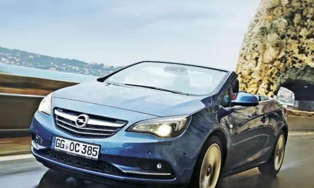 Niebieski Opel Cascada z opuszczonym dachem jedzie po mokrej drodze przy skalistym wybrzeżu.