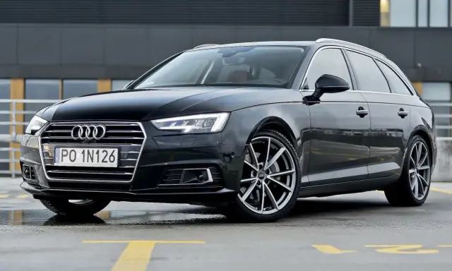 Czarny Audi A4 Avant, model b9, zaparkowany na mokrym parkingu z żółtymi liniami.
