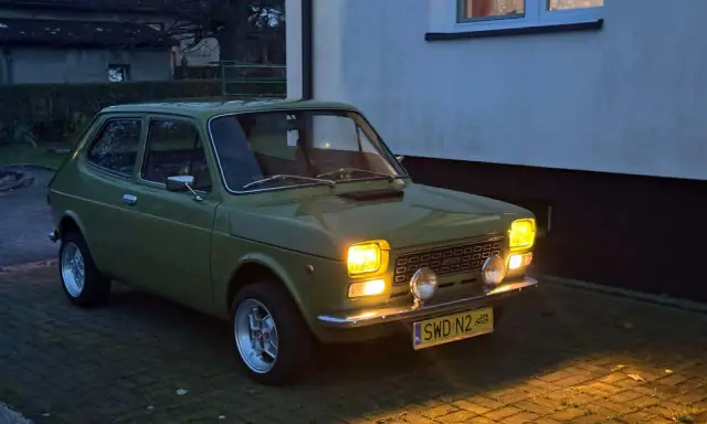 Zielony Fiat 127 z lat 70. stoi na podjeździe, jego światła rozświetlają zmierzch.