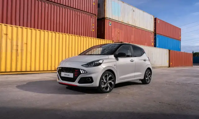 Szary Hyundai i10 N Line z czerwonymi akcentami na grillu i zderzaku, zaparkowany przed stosem kolorowych kontenerów. Sprawdź atrakcyjną hyundai i10 cena!