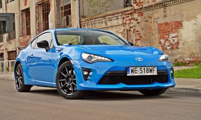 Błękitna Toyota GT86, znana też jako Toyota BRZ, zaparkowana przed zniszczonym budynkiem.
