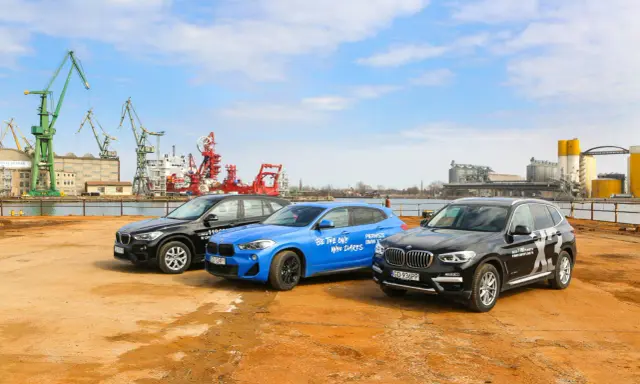 Trzy SUV-y BMW stoją na placu budowy, w tle portowe dźwigi i budynki.