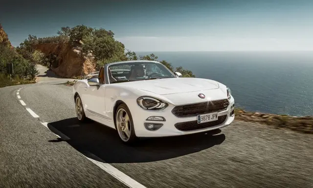 Biały Fiat 124 Spider sunie po krętej drodze nad morzem.