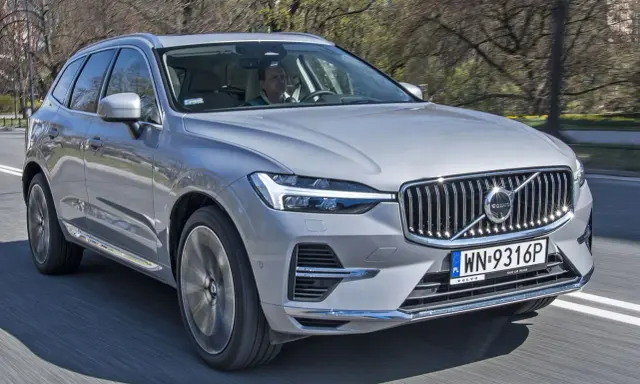 Srebrny Volvo XC60 2025 w ruchu, z kierowcą za kierownicą, na tle drzew.