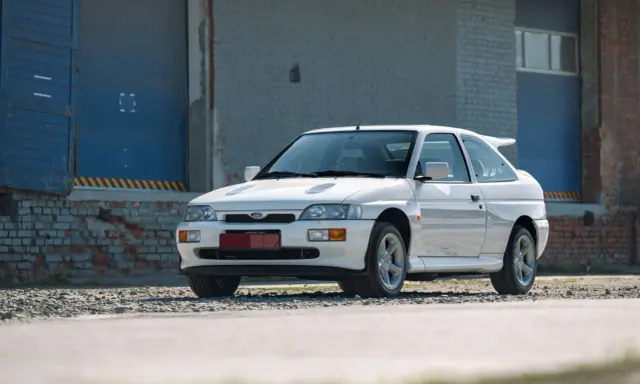 Biały Ford Escort Cosworth z charakterystycznym spojlerem, zaparkowany przed industrialnym budynkiem z niebieskimi drzwiami.