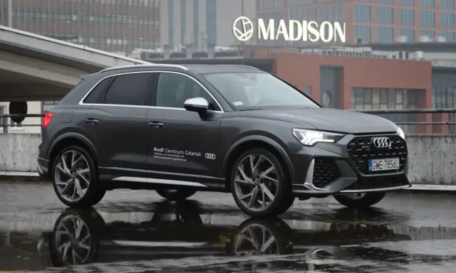 Szary Audi RS Q3 na mokrej nawierzchni, z odbiciem w kałuży. W tle budynek z napisem MADISON.
