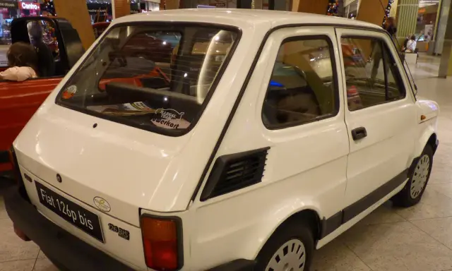 Biały Fiat 126p bis z naklejkami na tylnej szybie, stojący obok czerwonego samochodu.