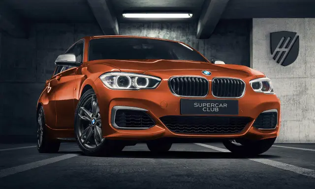 Pomarańczowe BMW 140i z tabliczką "SUPERCAR CLUB" stoi w garażu.