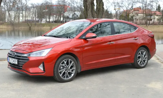 Czerwony Hyundai Elantra 2023 zaparkowany nad jeziorem, z drzewami i budynkami w tle.