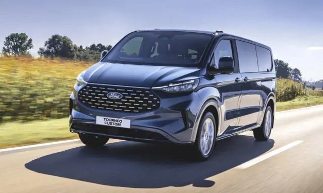 Ciemnoniebieski ford 7 osobowy Tourneo Custom pędzi po drodze wśród pól.