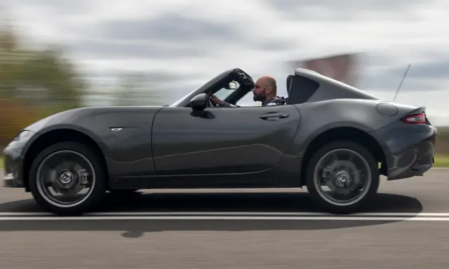 Mężczyzna w szarej kurtce prowadzi ciemnoszarą Mazdę Miata z opuszczonym dachem, jadąc po drodze.