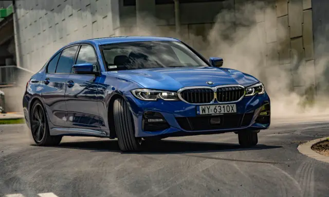 Niebieskie BMW 320i w zakręcie, unosząc tumany kurzu. Dynamiczna jazda na torze.