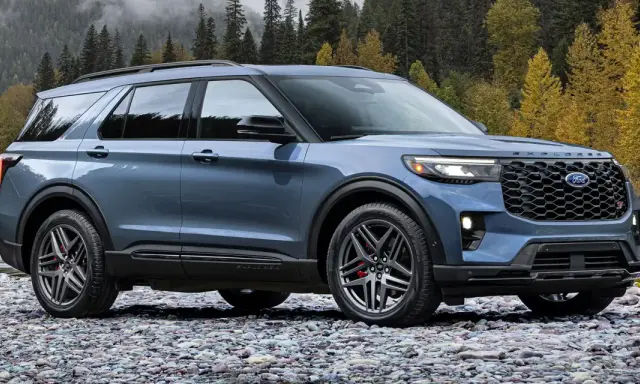 Niebieski Ford Explorer ST zaparkowany na kamienistej plaży, z jesiennym lasem w tle.