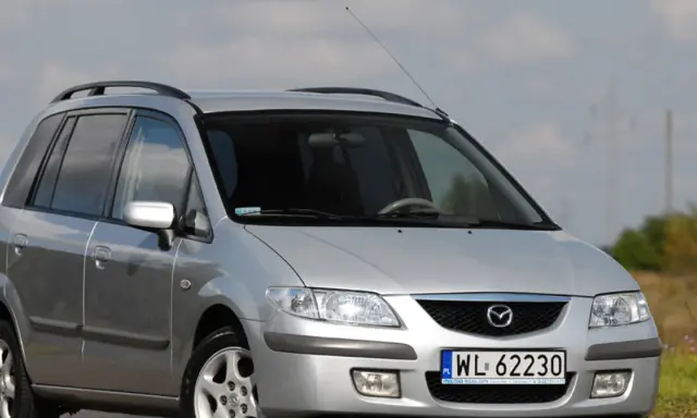 Srebrna Mazda Premacy z relingami dachowymi, zaparkowana na tle nieba i pól.