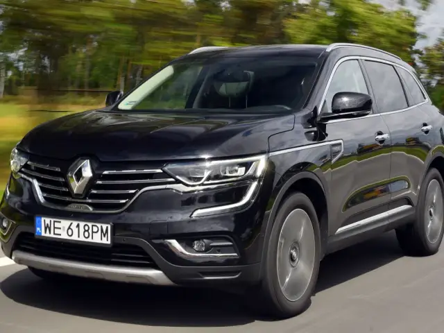 Czarny Renault Koleos 2.0 dCi pędzi po drodze, otoczony rozmytym zielonym krajobrazem.