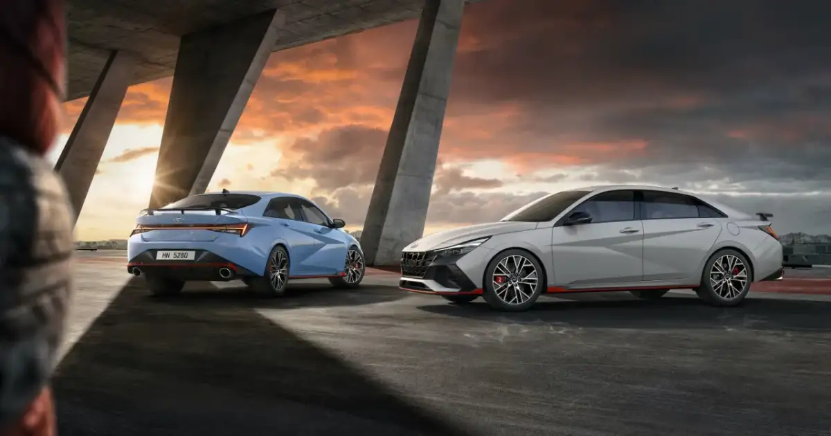 Dwa sportowe Hyundai Elantra N, jeden niebieski, drugi szary, stoją pod betonowym zadaszeniem o zachodzie słońca.