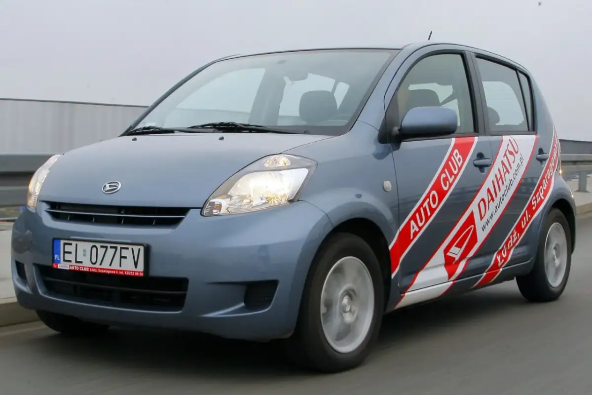 Niebieski Daihatsu Sirion z naklejkami "Auto Club Daihatsu" na drzwiach, poruszający się po drodze.