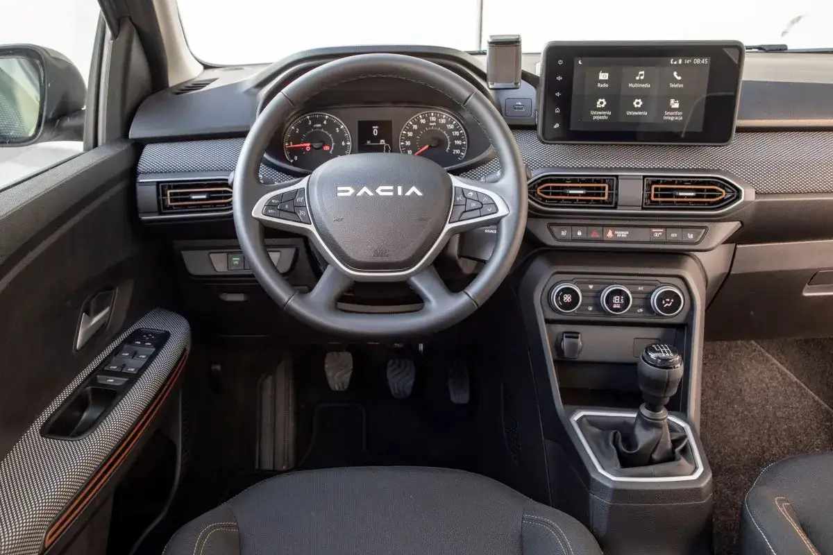 Wnętrze Dacii Sandero 2025: kierownica z przyciskami, ekran multimedialny, nawiewy i dźwignia zmiany biegów.
