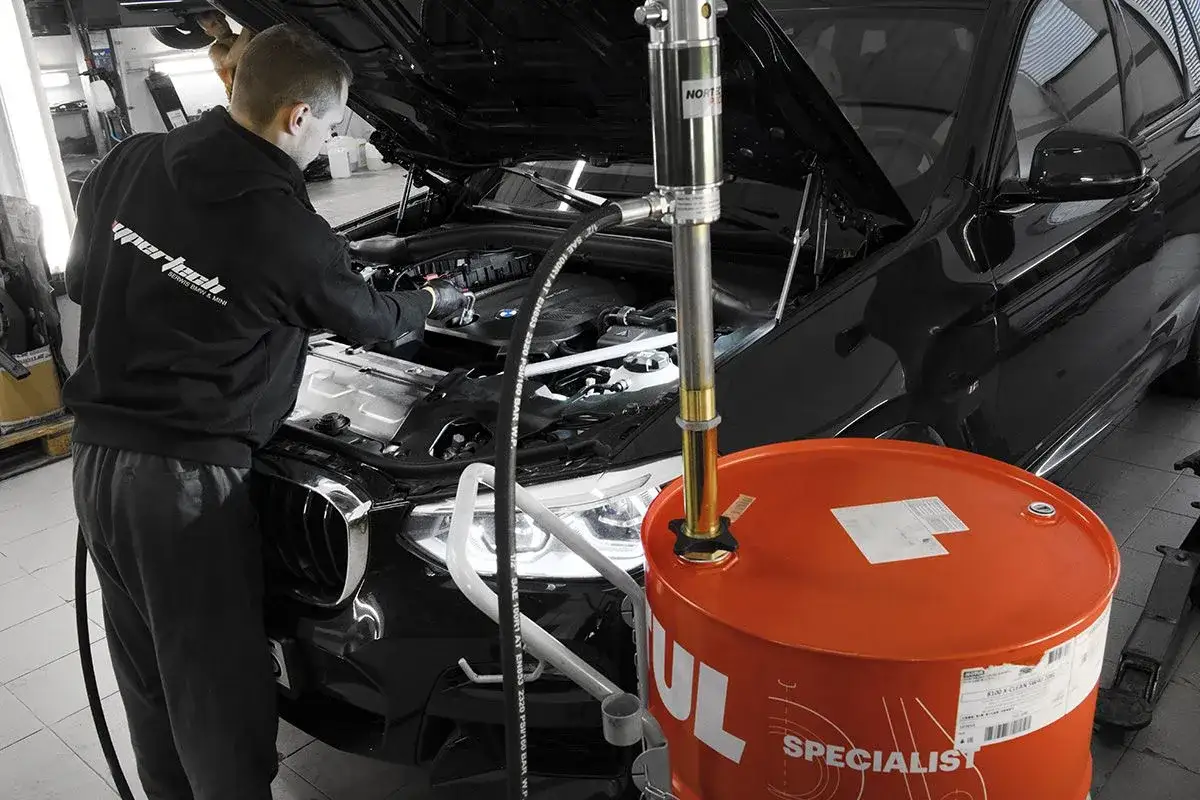 Mechanik wymienia olej w czarnym BMW. Obok stoi beczka z nowym olejem, sugerując, że co ile wymiana oleju jest kluczowa dla żywotności silnika.