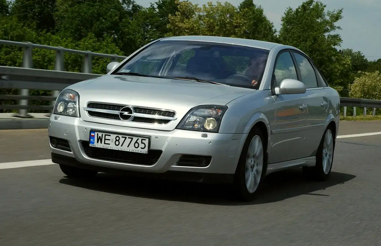 Srebrny Opel Vectra C GTS na drodze, z zielenią drzew w tle.