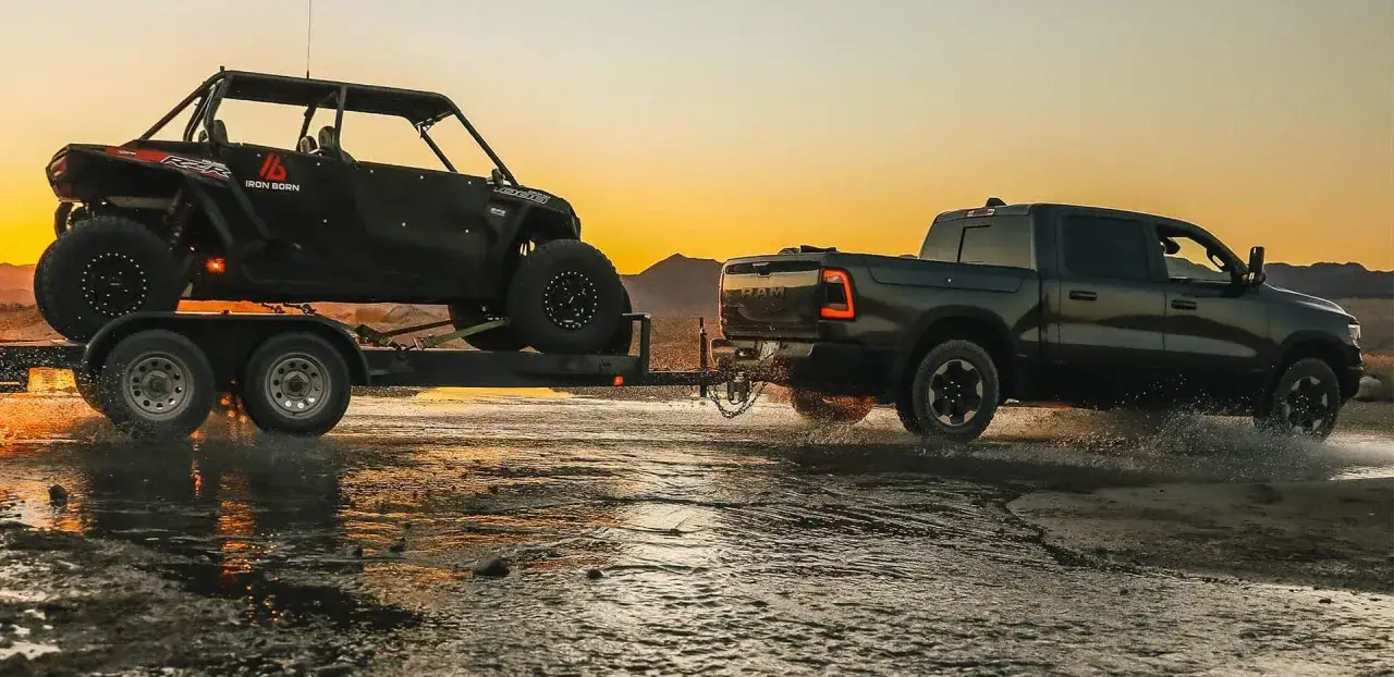Dodge Ram 1500 5.7 Hemi holuje przyczepę z quadem przez wodę o zachodzie słońca.