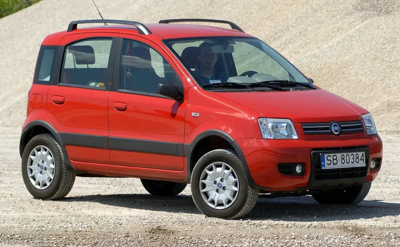 Czerwony Fiat Panda 2 z relingami dachowymi i czarnymi listwami, zaparkowany na żwirowym podłożu. Kierowca widoczny w kabinie.