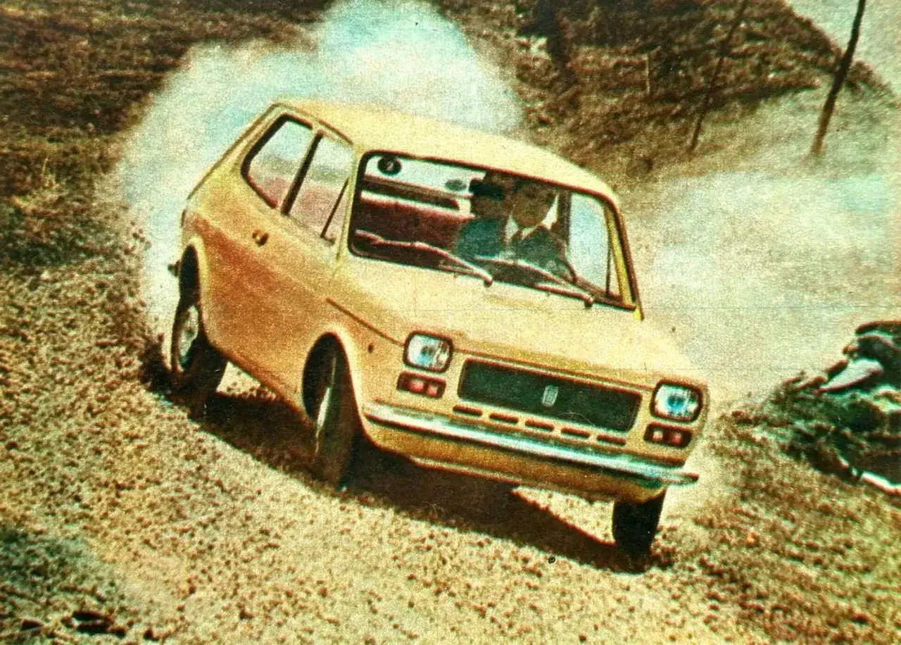 Żółty Fiat 127 z dwoma osobami w środku, pokonujący zakręt na szutrowej drodze, wzbijając tumany kurzu.