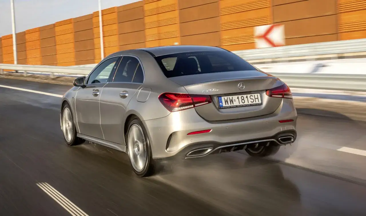 Srebrny Mercedes A250e pędzi po mokrej drodze, rozpryskując wodę. Pomarańczowa ściana dźwiękochłonna w tle.