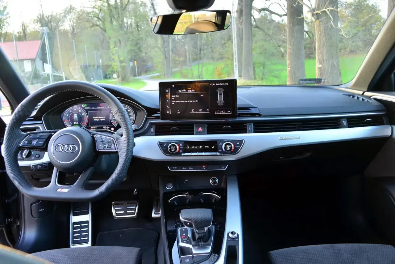 Wnętrze Audi z kierownicą, ekranem multimedialnym i panelem klimatyzacji. Widok na drogę wśród drzew.